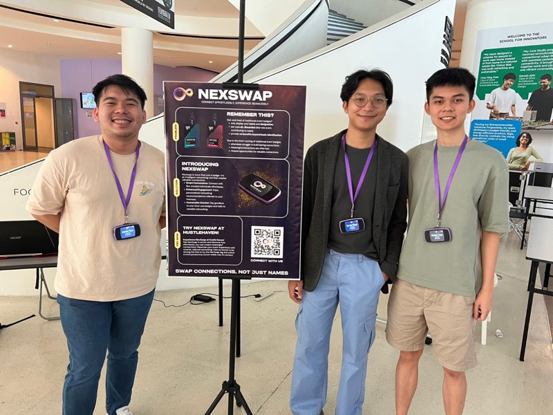 Three of the NexSwap team wearing badges beside the NexSwap poster at Hustle Haven